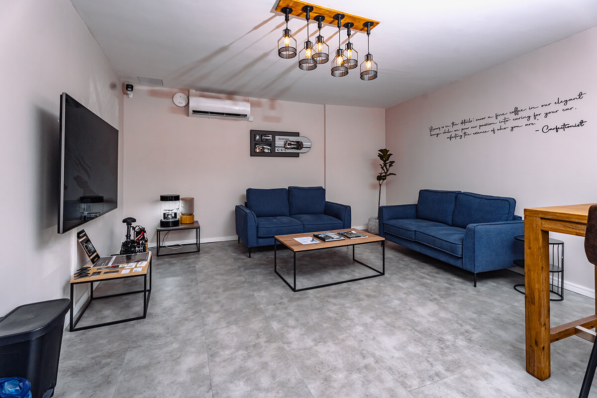 Modern car detailing lounge with two blue sofas, coffee tables, large wall-mounted TV, and industrial ceiling lights in a minimalist room.